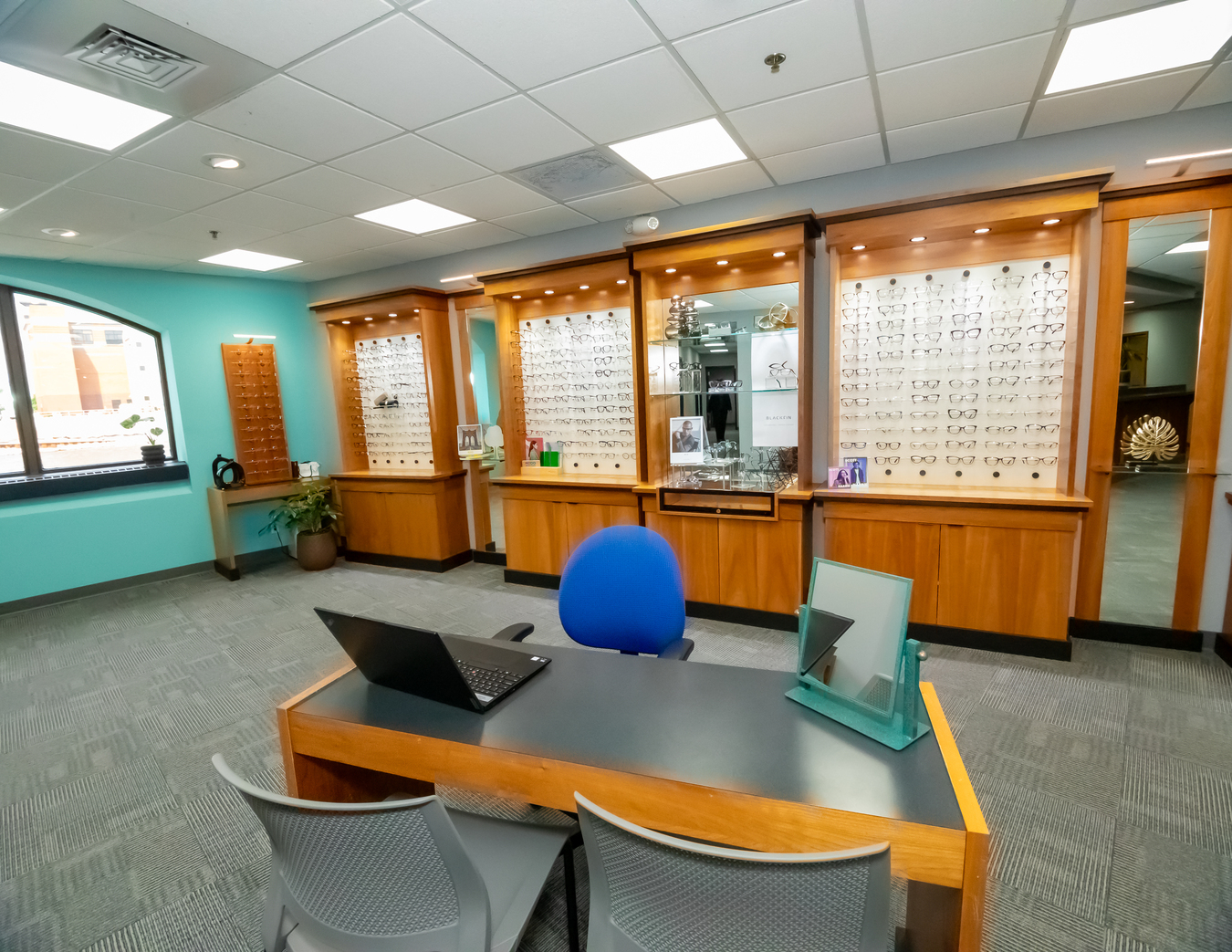 An optometrist helps a patient try on glasses while sitting at a desk with a computer displaying eyewear options - Carlson Eye Care